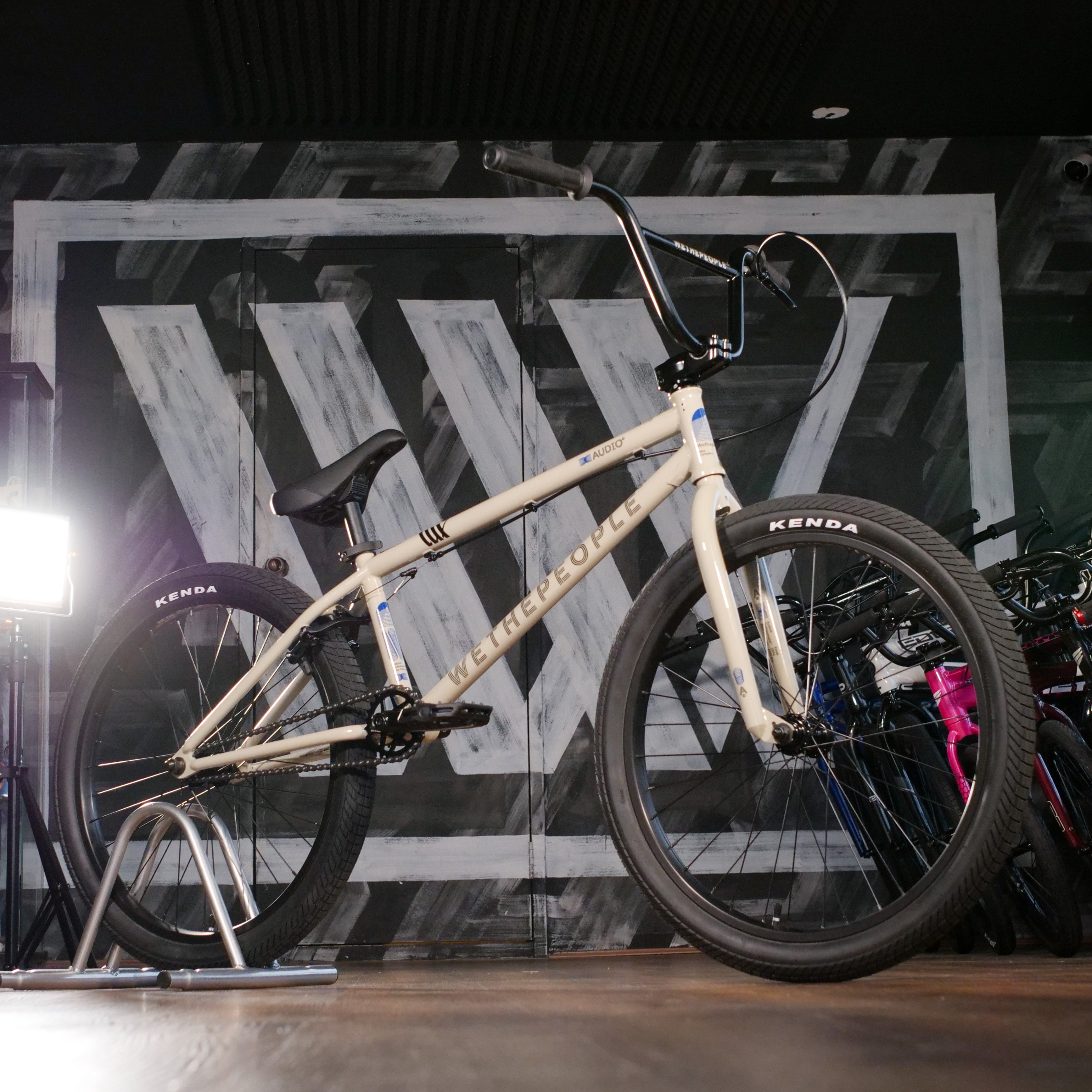 The Wethepeople Audio 24 Inch Bike, featuring a durable beige chromoly frame and Kenda tires, is displayed indoors on a bike stand against a wall with a large geometric "W" design.