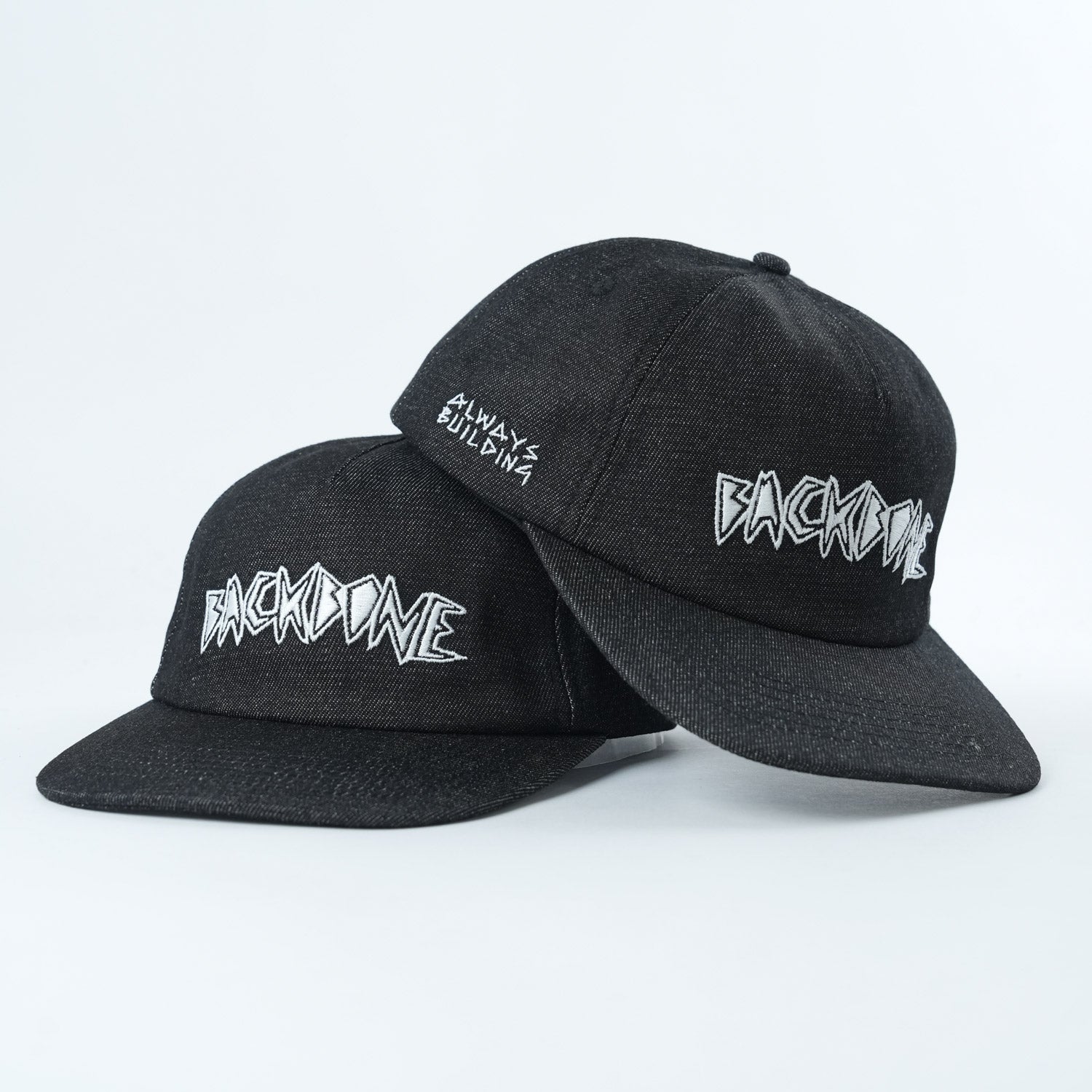 Two black Backbone Always Building Hats—a BMX snapback and an unstructured 5 panel—feature "BACKBONE" embroidered on the front and "ALWAYS BUILDING" on the side, shown against a plain white background.