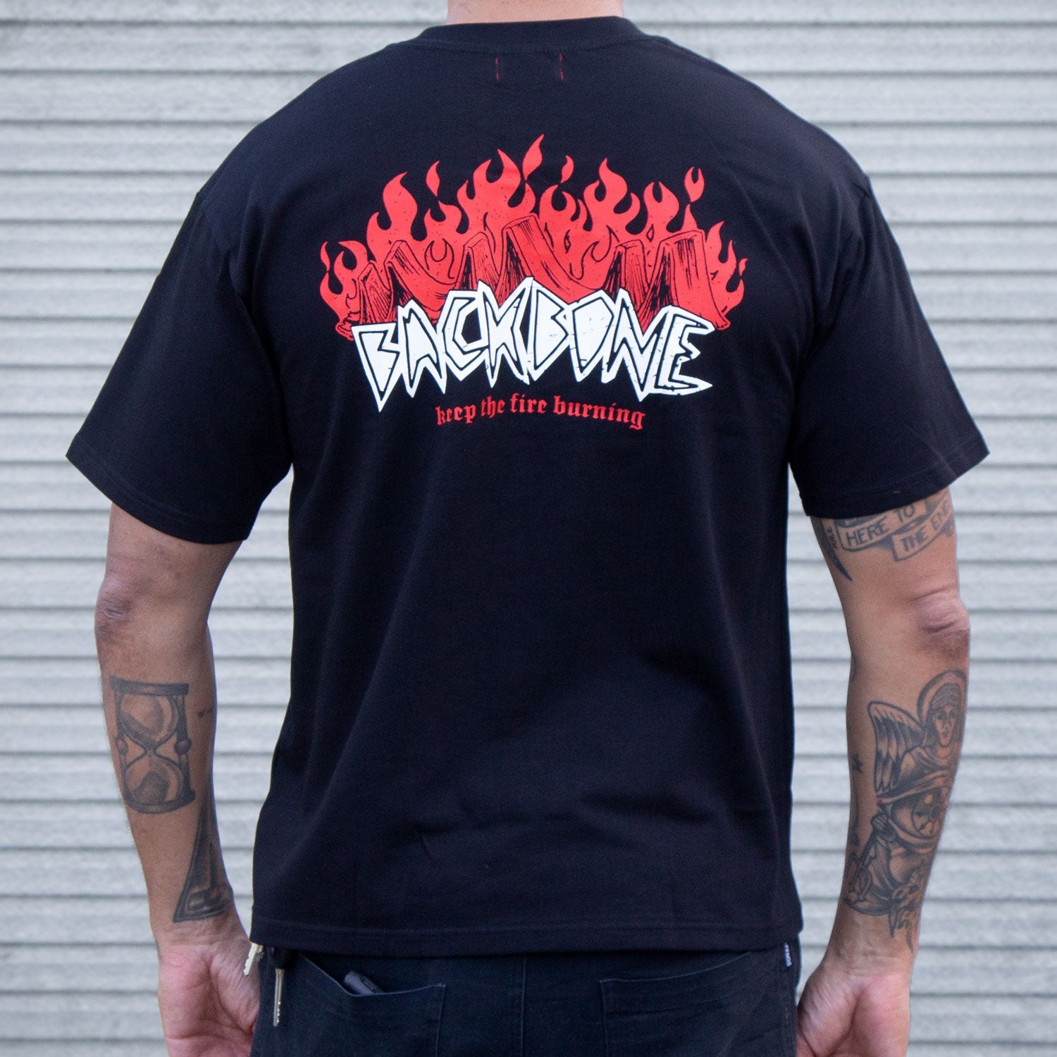 Sporting the Backbone Fire Burning T-Shirt—premium black cotton with red flames, "BACKBONE" in white, and "keep the fire burning" in red on the back—a tattooed wearer channels Aussie BMX vibes.