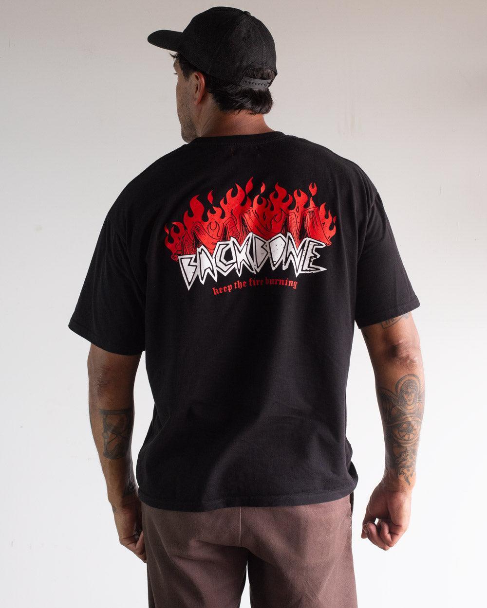 A man faces away, wearing the Backbone Fire Burning T-Shirt with red flame graphics and "BACKBONE keep the fire burning" text on the back, styled with brown pants, a black cap, and embodying classic Aussie BMX attitude.