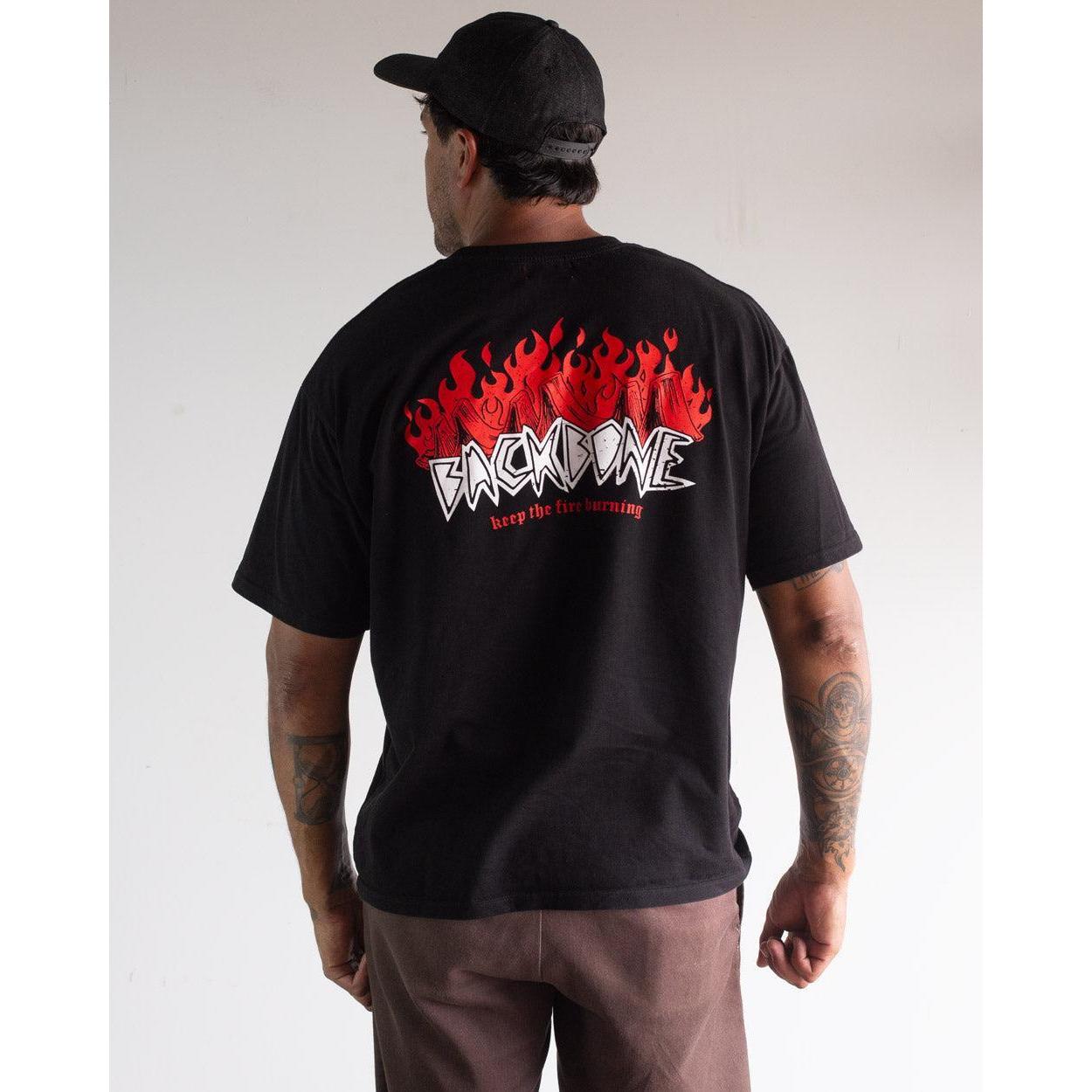 A man faces away, wearing the Backbone Fire Burning T-Shirt with red flame graphics and "BACKBONE keep the fire burning" text on the back, styled with brown pants, a black cap, and embodying classic Aussie BMX attitude.