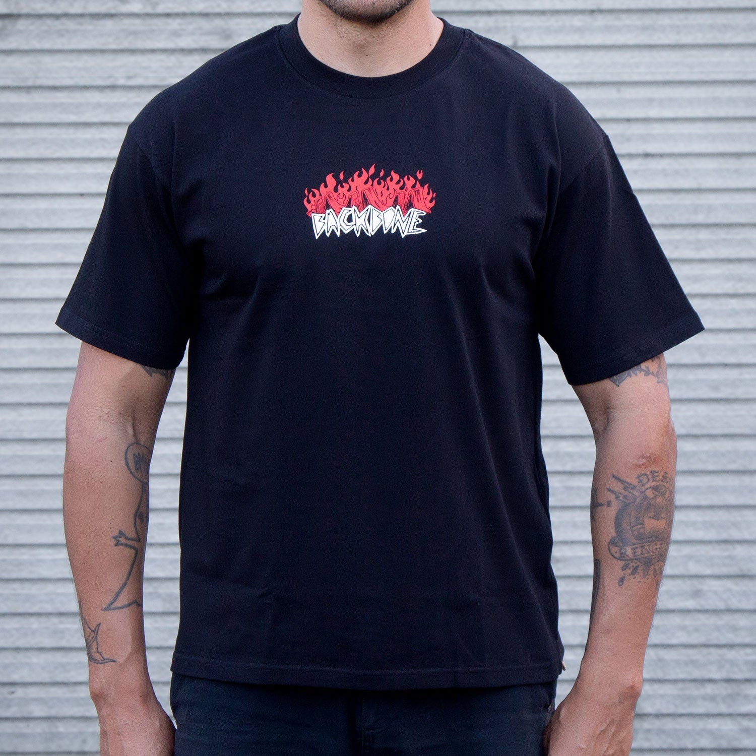 A person wears the Backbone Fire Burning T-Shirt, featuring red flames and bold white "BACKWAVE" text on premium heavyweight cotton, standing before a corrugated metal wall.