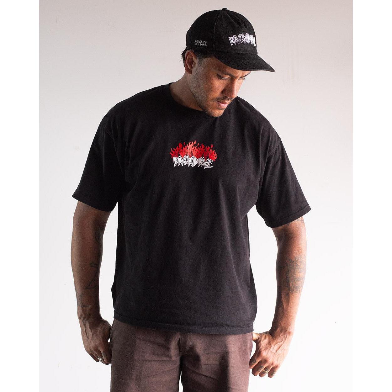 Man wearing the Backbone Fire Burning T-Shirt—a premium cotton, heavyweight tee featuring a red and white "BROOKLYN" graphic—paired with a matching black cap, standing against a plain background.
