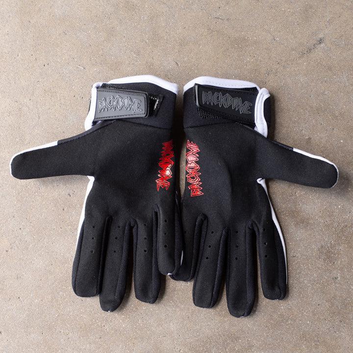 The Backbone Gnar Drip Adult Gloves, black with white edges and red palms adorned with lettering, made from lightweight spandex, are laid flat on a concrete surface.