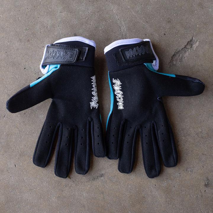 A pair of Backbone Gnar Drip Adult Gloves made from lightweight spandex, in black and teal with Velcro straps and white stitching, lies on a concrete surface.
