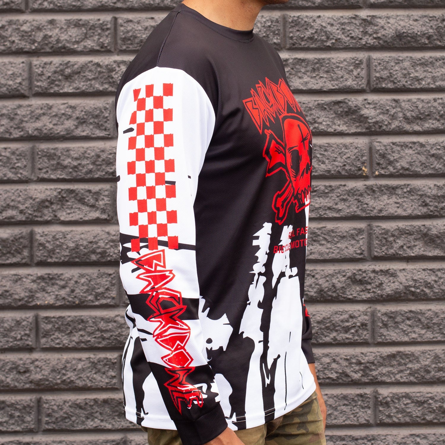 A man in the Backbone Real Fast Race Jersey / Gnar Drip (Adult), featuring red and white designs with a skull and checkered pattern, stands against a gray brick wall.