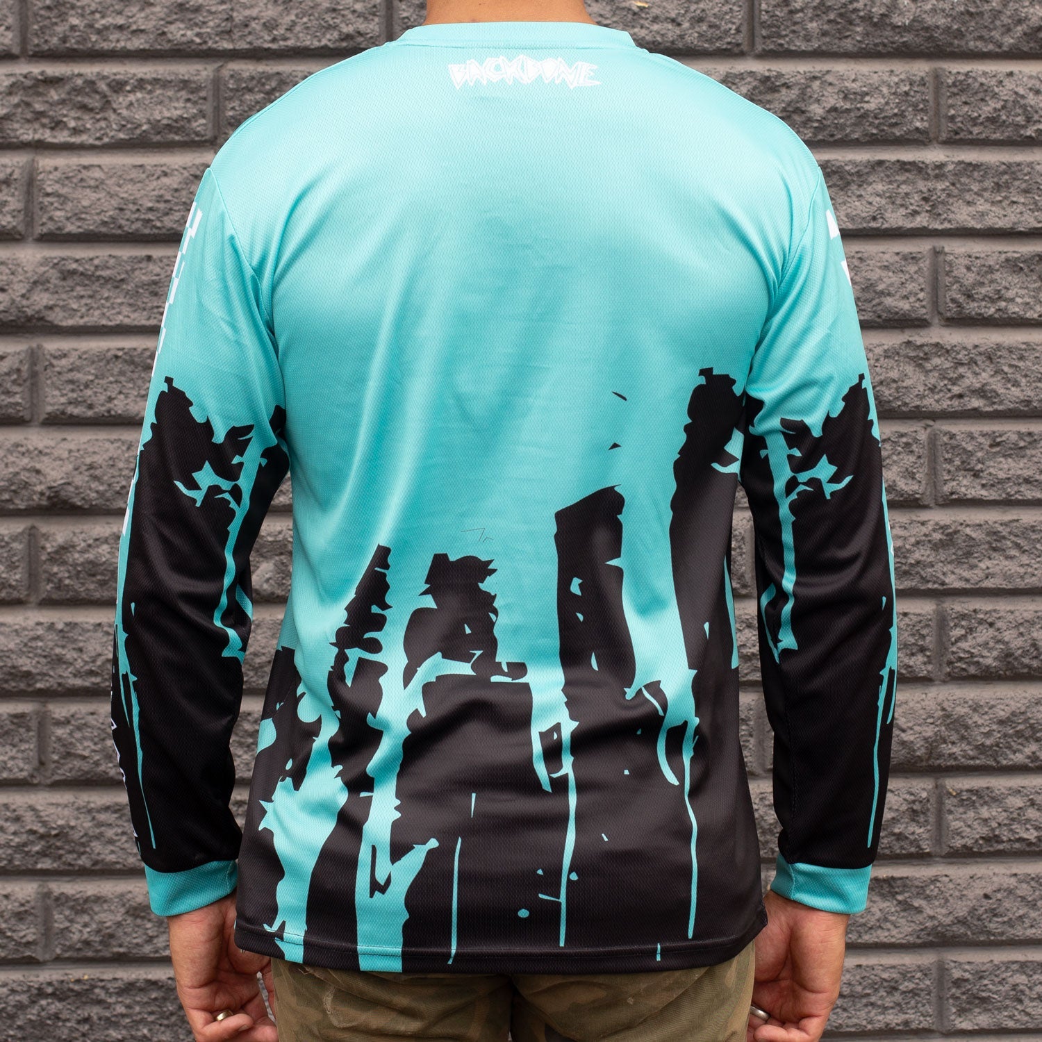 A person stands in front of a gray brick wall wearing the "Backbone Real Fast Race Jersey / Gnar Drip (Youth)," a custom sublimated turquoise and black long-sleeve shirt made from 160 GSM polyester, featuring abstract tree-like designs on the back.