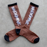 The Backbone Crew Socks are buttery soft, brown mid-shin socks with black toes, heels, and cuffs, featuring a white zigzag design along the sides and displayed on a grey surface.