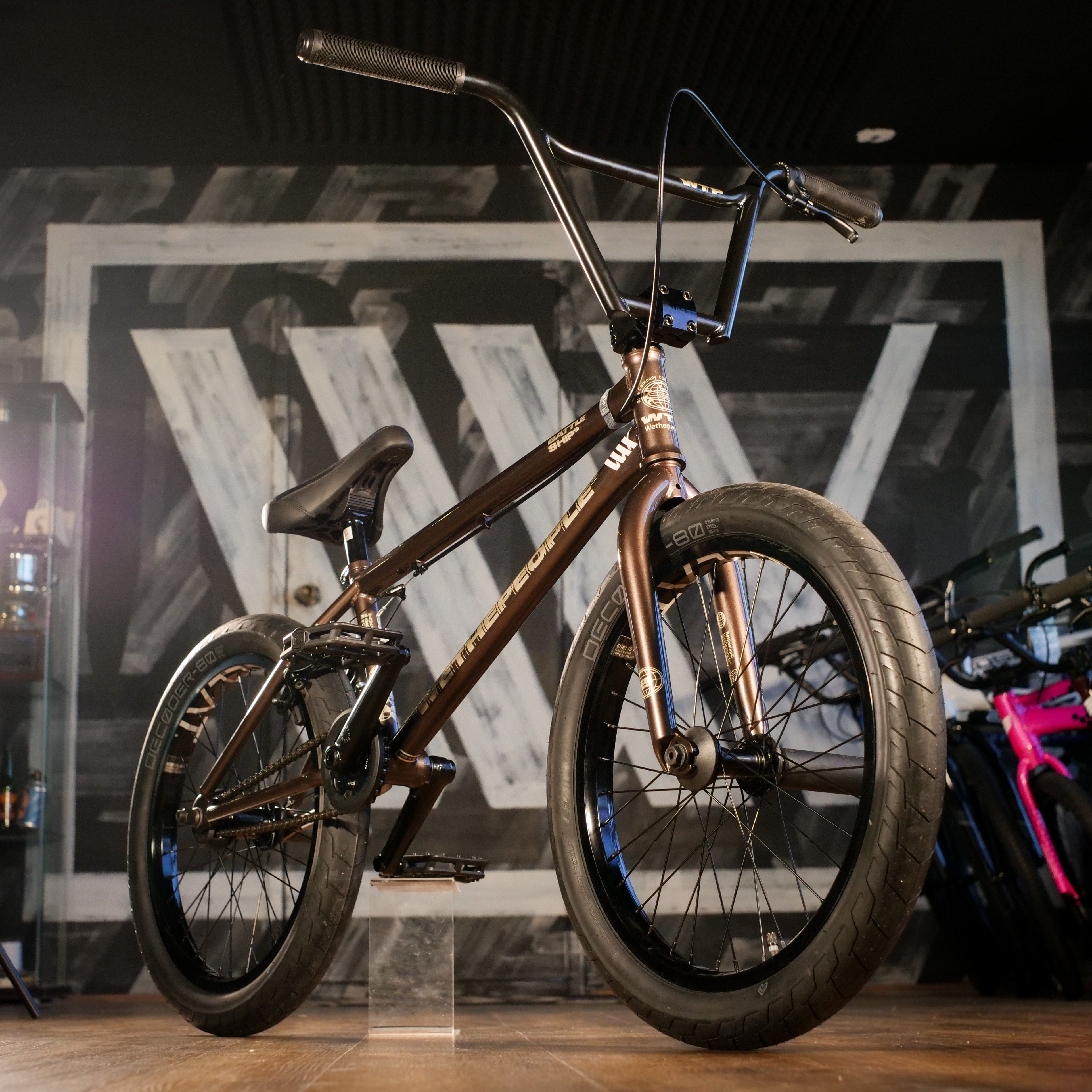 The Wethepeople Battleship 20 Inch Bike, with a bronze 4130 Chromoly frame, black handlebars, and tires, is displayed indoors on a wooden floor with a painted mural in the background.