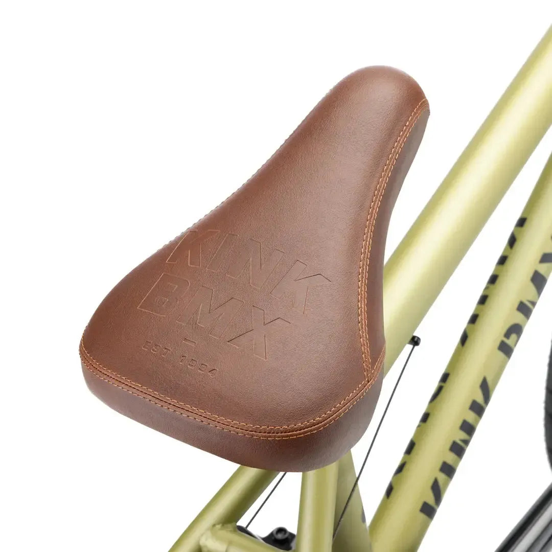 A close-up of a brown leather BMX seat embossed with "KINK BMX" and "EST 1994," mounted on the gold Kink Gap XL 20 Inch Bike (2026) frame made from strong 4130 chromoly.
