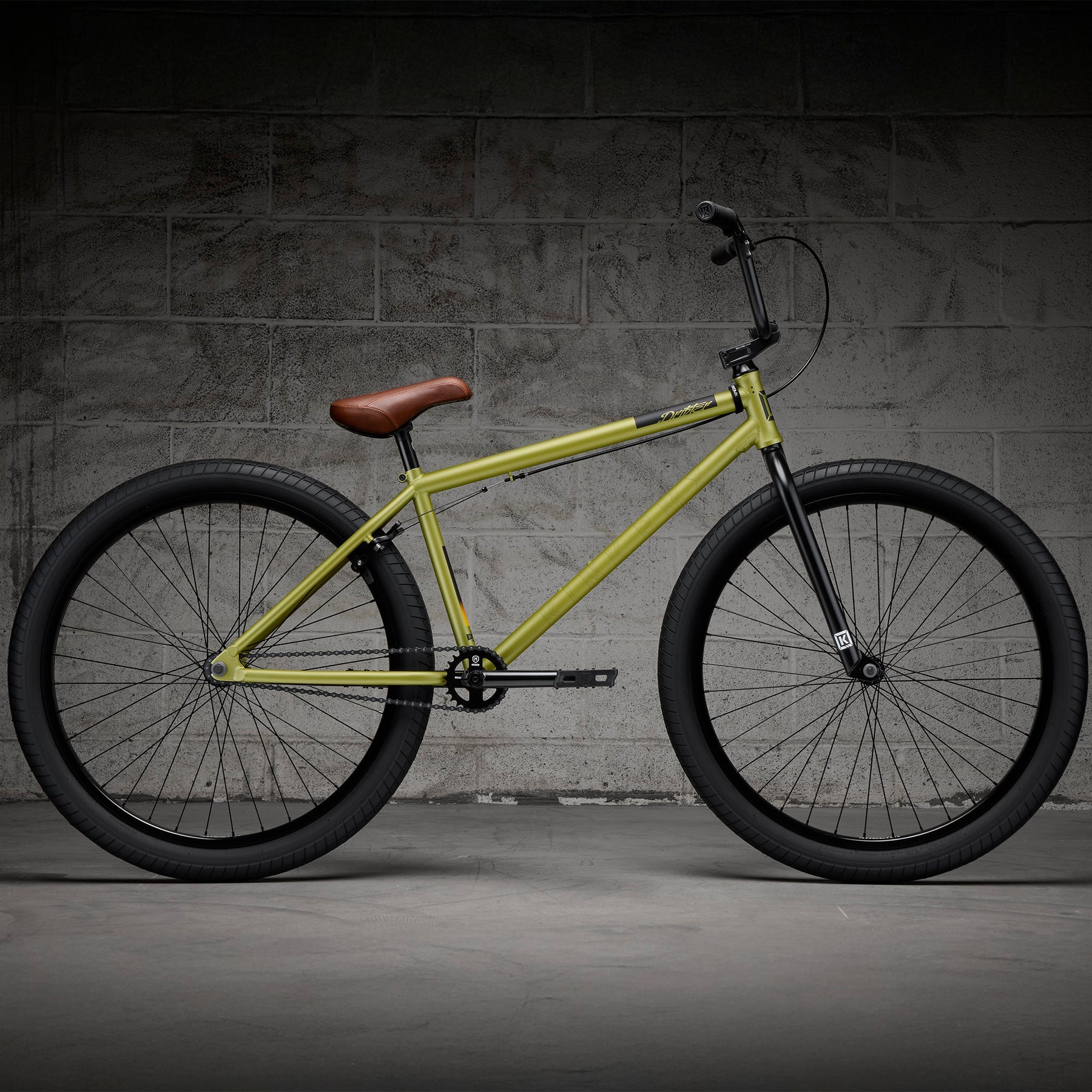 A Kink Drifter 26 inch Bike (2027) in yellow, featuring black wheels and a brown saddle, is shown sideways against a gray concrete block wall.