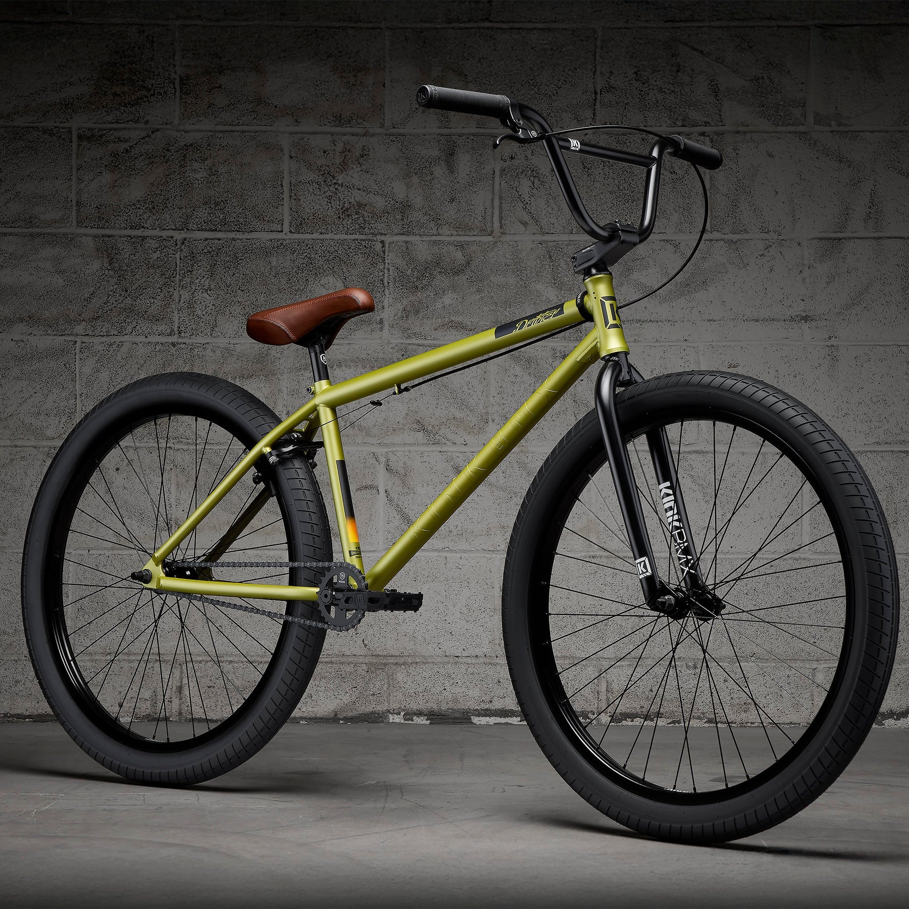 A Kink Drifter 26 inch Bike (2027) featuring a yellow chromoly frame, black tires, and a brown saddle is parked on a concrete floor in front of a gray brick wall.