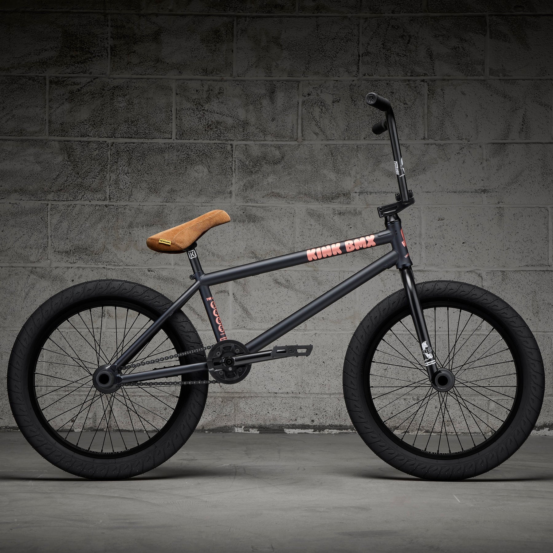 A Kink Williams 20 Inch Bike (2027) with thick tires, a tan seat, and a durable black chromoly frame is set against a gray concrete wall background.