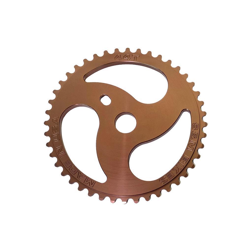 The S&M Chainsaw Sprocket is a copper-colored, CNC machined 44-tooth bicycle chainring with three large cut-outs and "MADE IN USA" and "S&M BIKES" engravings, styled like a chainsaw sprocket for both durability and unique looks.