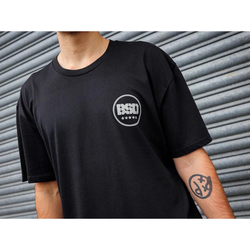 A person in a BSD Fully Roasted T-Shirt with a circular PSU logo stands against corrugated metal, showcasing a forearm tattoo that suggests a love for double shots of caffeine.