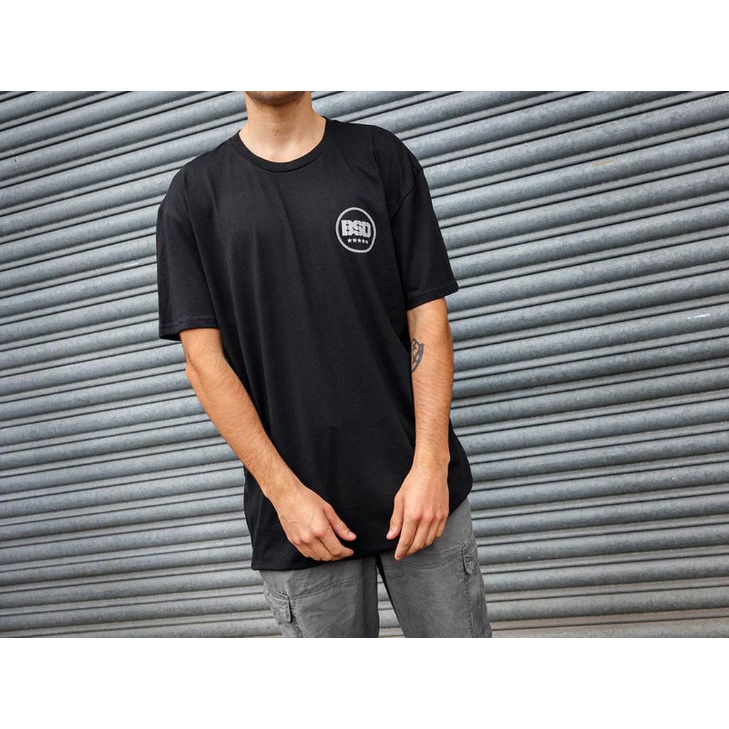Clad in a black BSD Fully Roasted T-Shirt and gray cargo shorts, the person stands against a corrugated metal backdrop, holding a double shot of coffee for an extra kick.