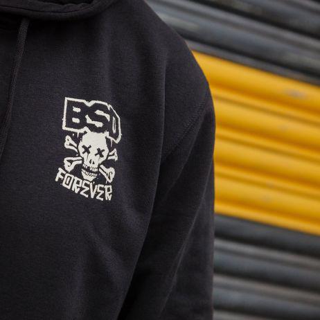 Close-up of a BSD More Speed Hoodie, a black garment with a skull and crossbones design labeled "BSD Forever," styled against gray and yellow metal shutters.
