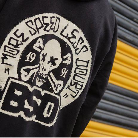 Close-up of the BSD More Speed Hoodie, a stylish black sweatshirt with a skull and crossbones graphic. "MORE SPEED LESS DOUBT" is prominently displayed along with "1991" and "BSD," set against yellow and gray shutters.