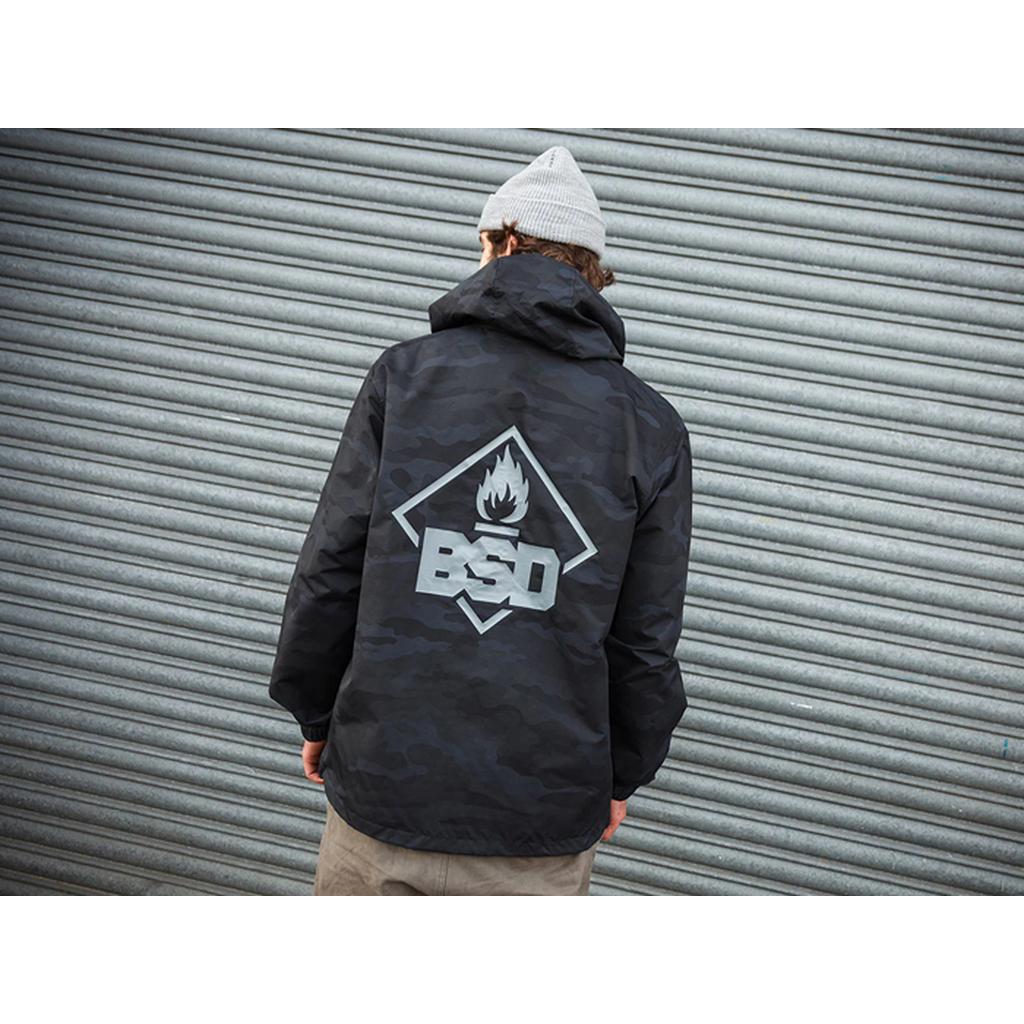 A person wearing the BSD Roastin Jacket, showcasing a black camo with a flame logo and "BSD" on the back, confidently poses in front of a corrugated metal wall while sporting a gray beanie.