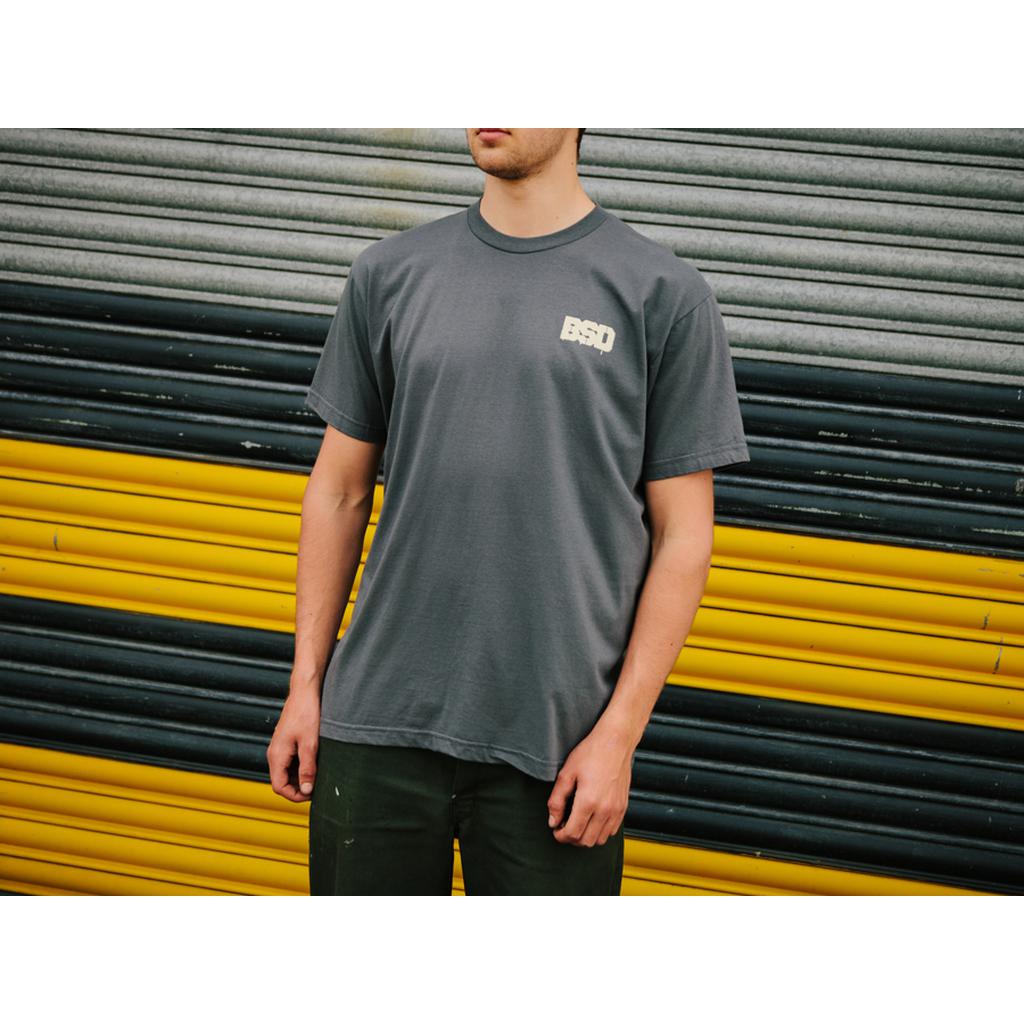 A person in a BSD Spillage T-Shirt (Grey) stands before a closed metal shutter striped in yellow and black.