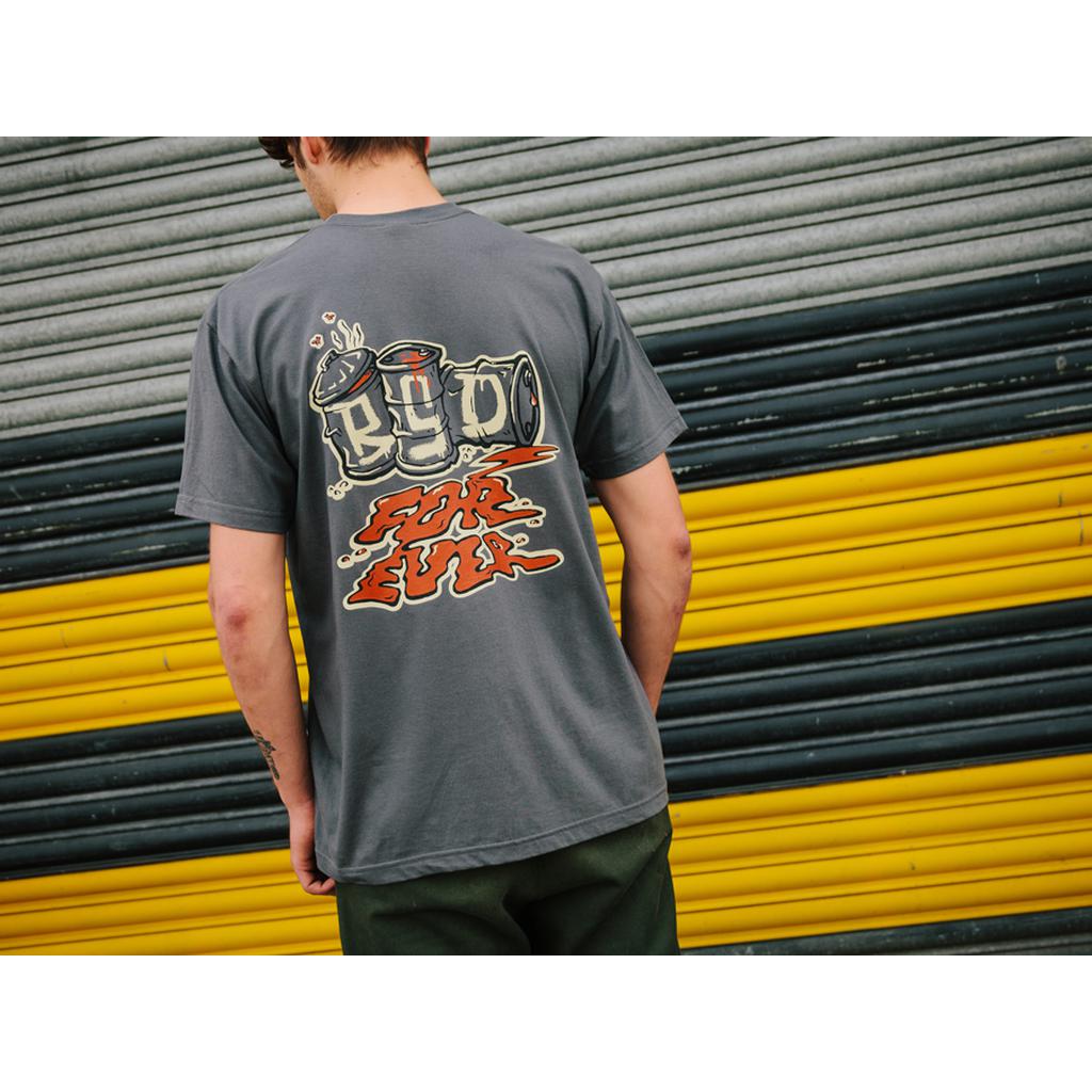 A person in a BSD Spillage T-Shirt (Grey), featuring a graffiti-style design, stands before a striped metal backdrop, complemented by subtle embroidered BSD tags.