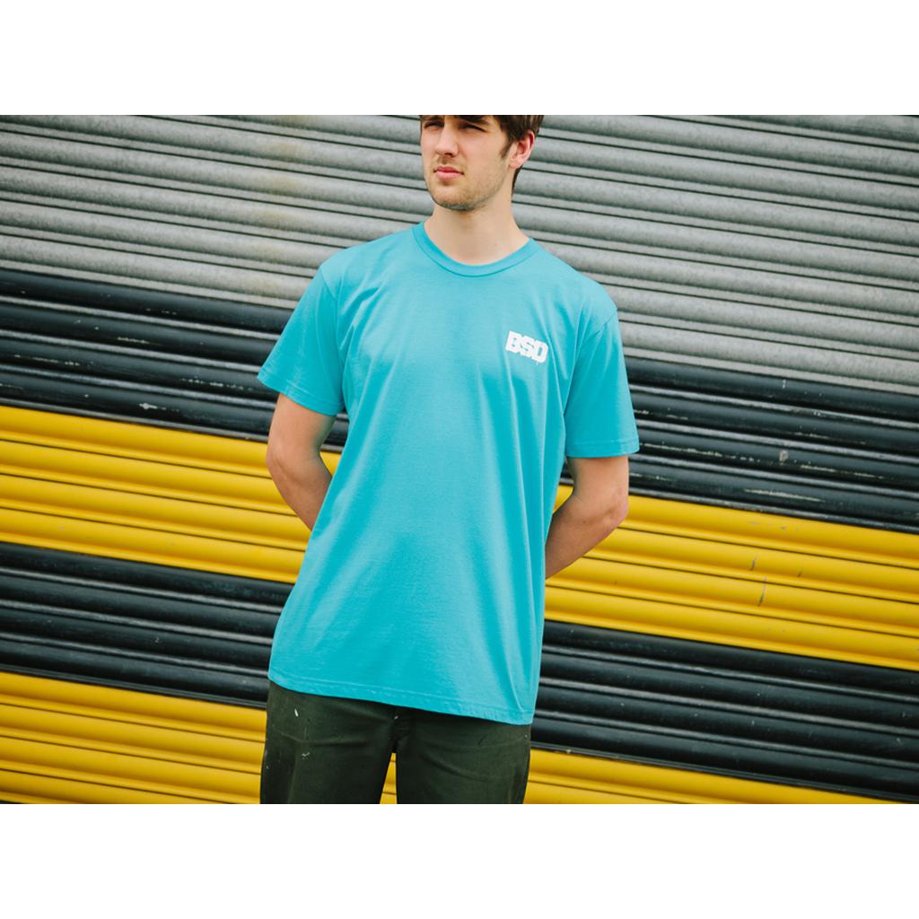 A person in a blue BSD Spillage T-Shirt, crafted from lightweight cotton for superior comfort, stands before a corrugated shutter decorated with black, yellow, and gray stripes, gazing to the side.