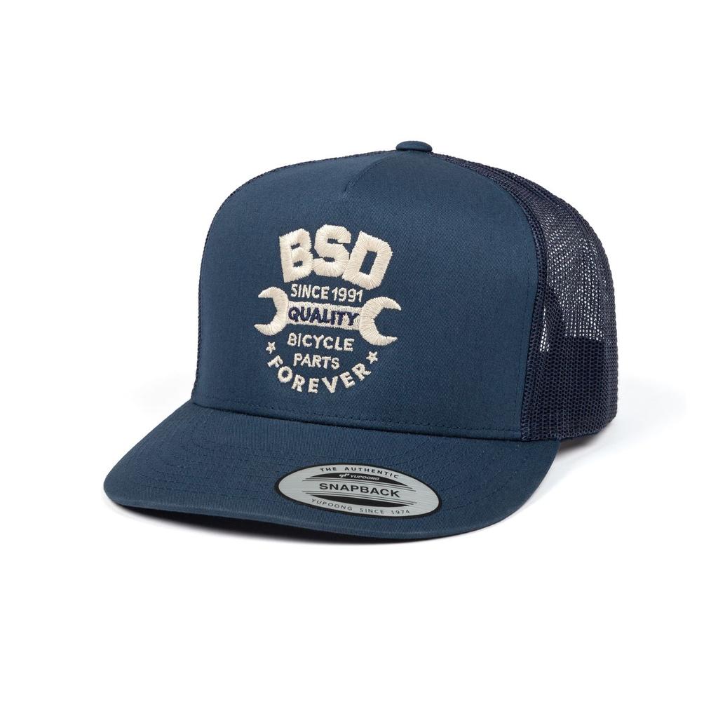 The BSD Workshop Trucker Cap is a navy blue mesh cap with "BSD Since 1991 Quality Bicycle Parts Forever" and wrench graphics embroidered on the front—ideal for bike cap collectors.