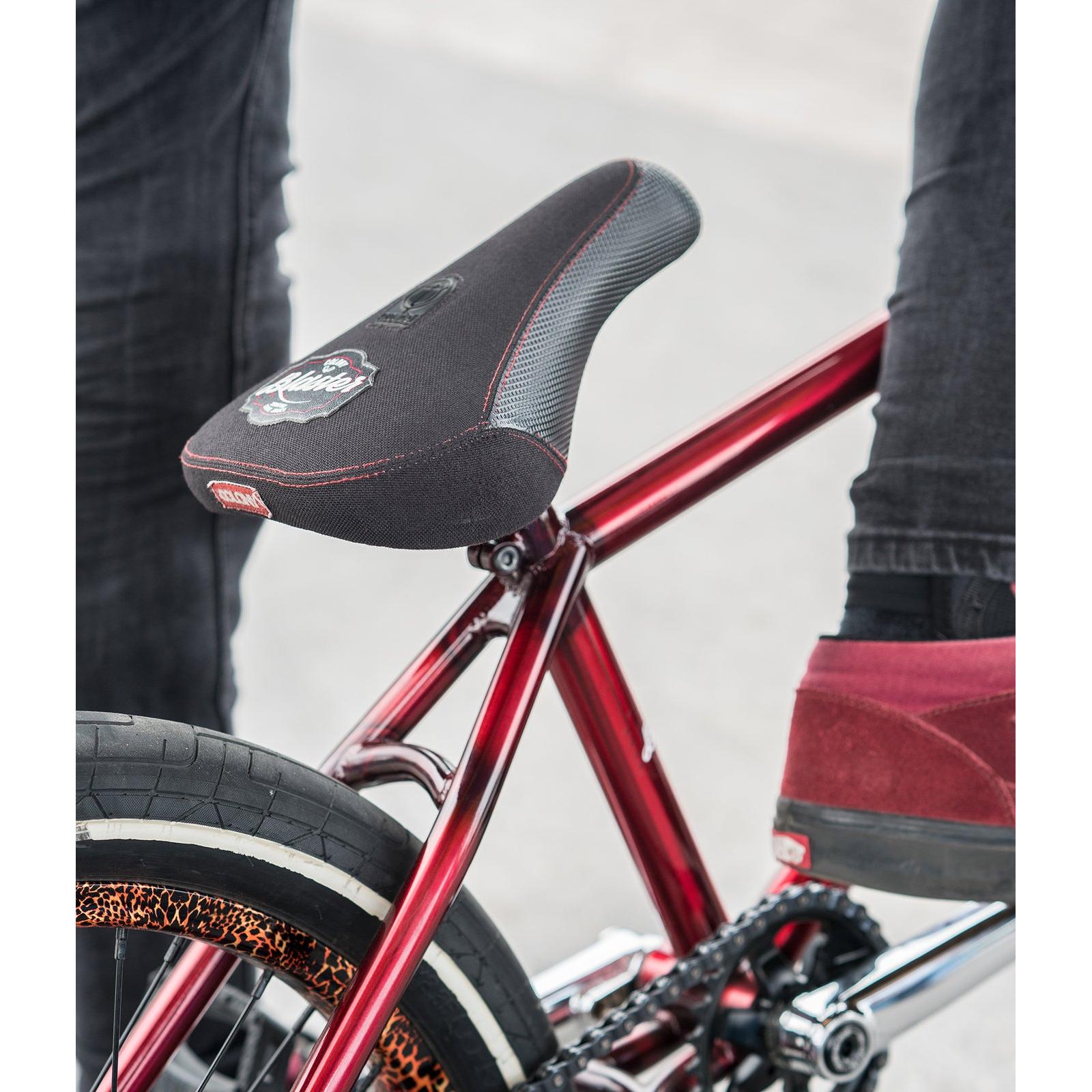 Close-up of a red bicycle featuring the Colony Blaster Fat Pivotal Seat, partially visible rear tire, and a person in red shoes standing nearby.