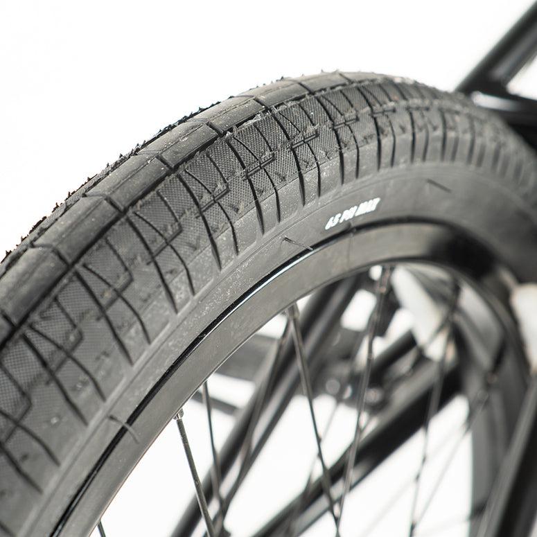 Close-up of a Colony Sweet Tooth Pro Elite 18 Inch BMX tire with a textured tread and visible spokes. Marked "65 PSI MAX," the tire rests against a blurred background suggesting motion and adventure.
