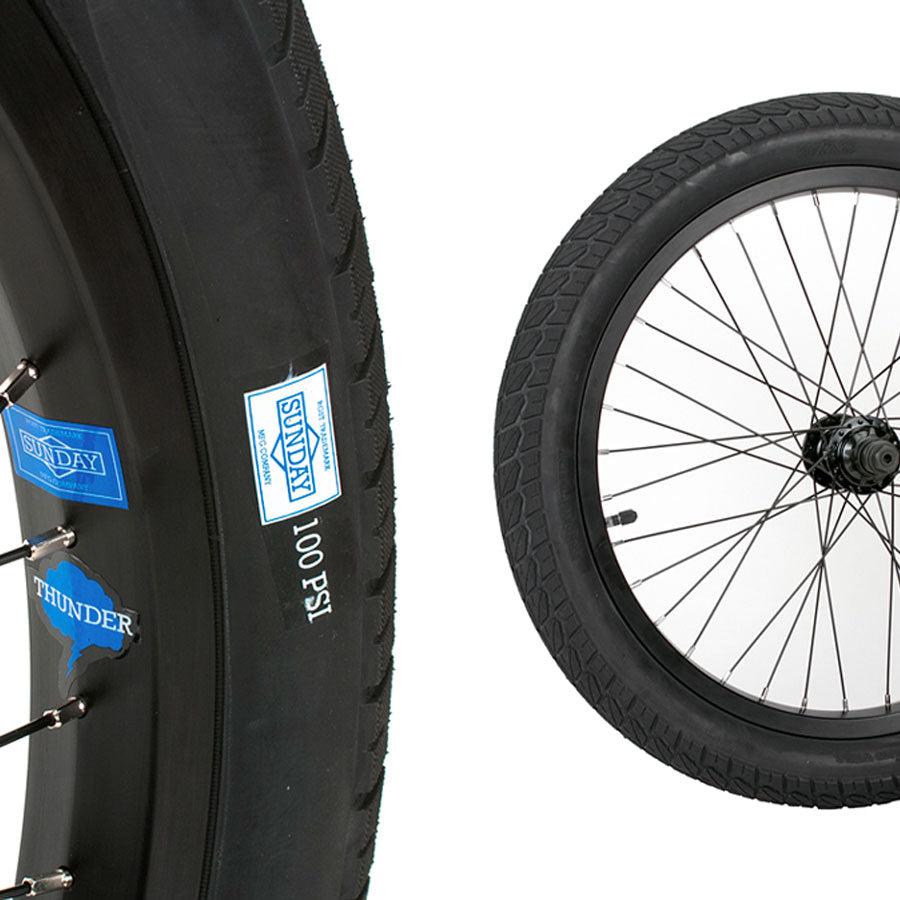 Close-up of two bicycle wheels with thick, grippy tires; one featuring a blue "Sunday" label and a "Thunder" sticker, indicating a 100 PSI rating. Ideal for beginner riders, these Sunday Current 18 Inch Tyres ensure excellent traction and stability.