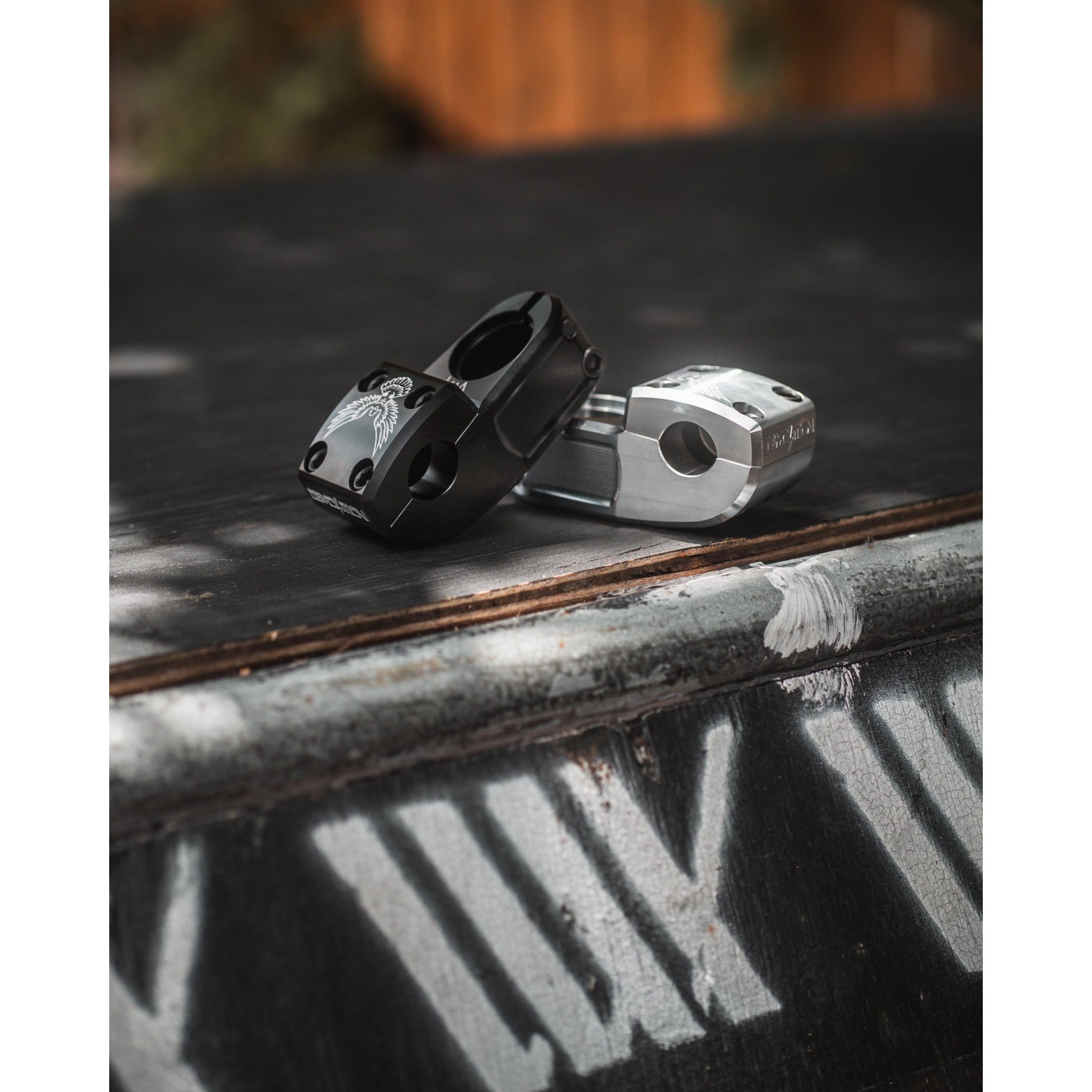 Two metal BMX bike stems, including the Demolition Josh Dove Stem, are displayed on a rough surface. One is black, and the other is silver, with stencil-like text in the background enhancing their rugged appeal.