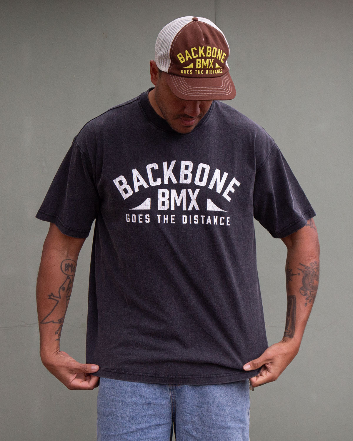 A man wearing the Backbone Distance Vintage T-Shirt and a cap stands against a plain background, looking down while holding the bottom of his shirt.