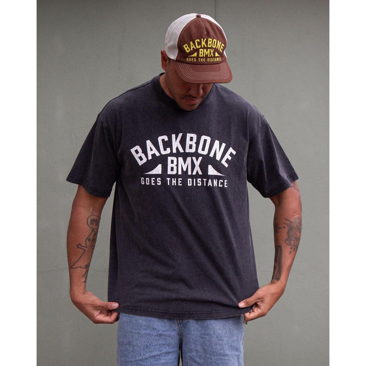 A man wearing the Backbone Distance Vintage T-Shirt and a cap stands against a plain background, looking down while holding the bottom of his shirt.