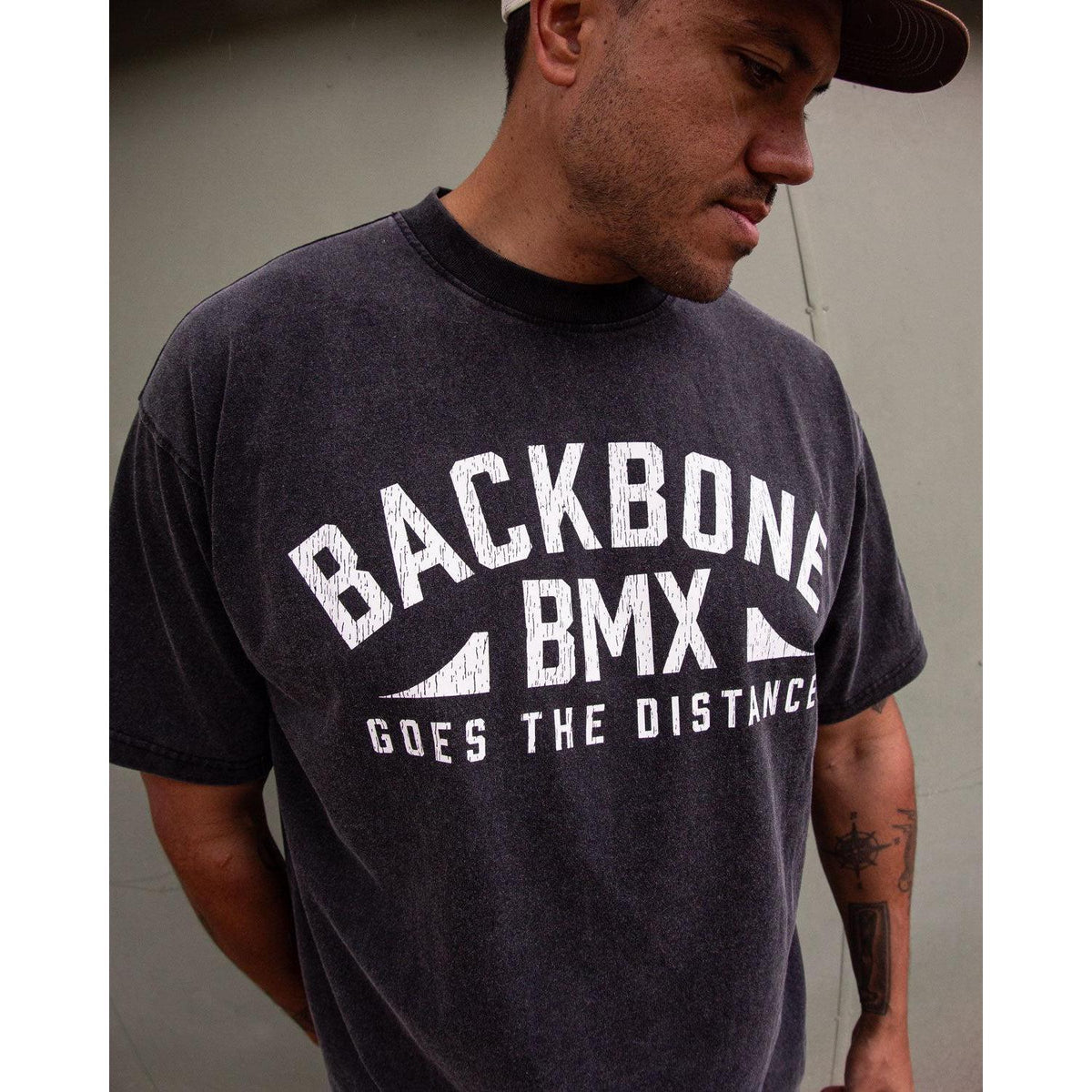Wearing the Backbone Distance Vintage T-Shirt featuring the text “BACKBONE BMX GOES THE DISTANCE,” a person stands against a plain wall, gazing down and to the side.