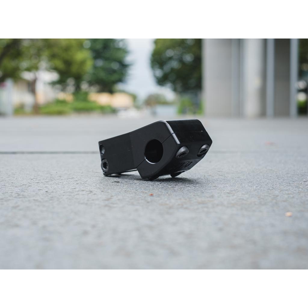The Eclat 1023 Stem, a CNC machined black metal bracket, rests on a concrete surface outdoors with blurred trees and buildings creating a picturesque backdrop.