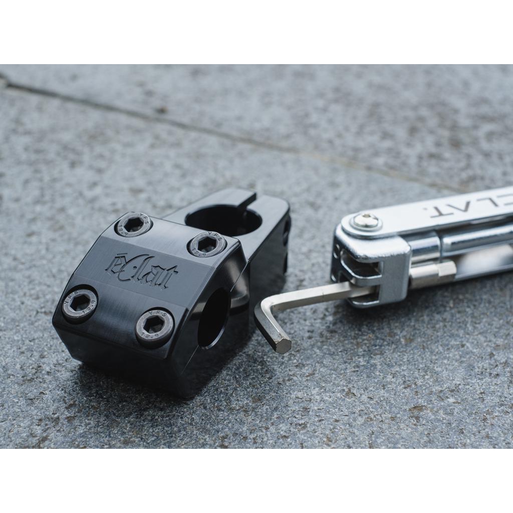 The Eclat 1023 Stem, featuring a black mid-school style with a logo, is displayed on a gray surface next to a multi-tool with an Allen key extended.