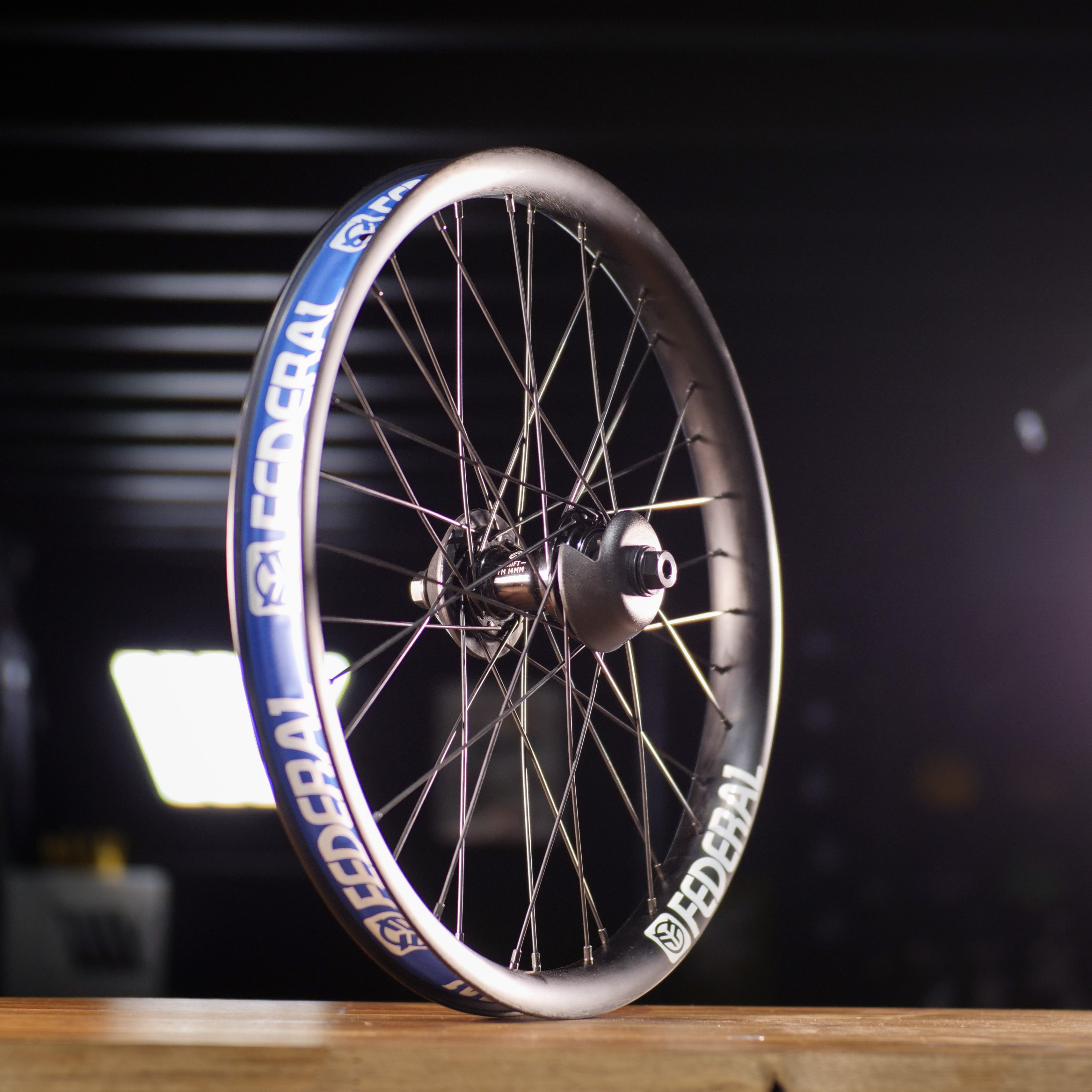 A Federal X Eclat FM Shift Hybrid Custom Wheel with black spokes, a blue rim, and a hybrid hub stands upright on a wooden surface in a dimly lit room.