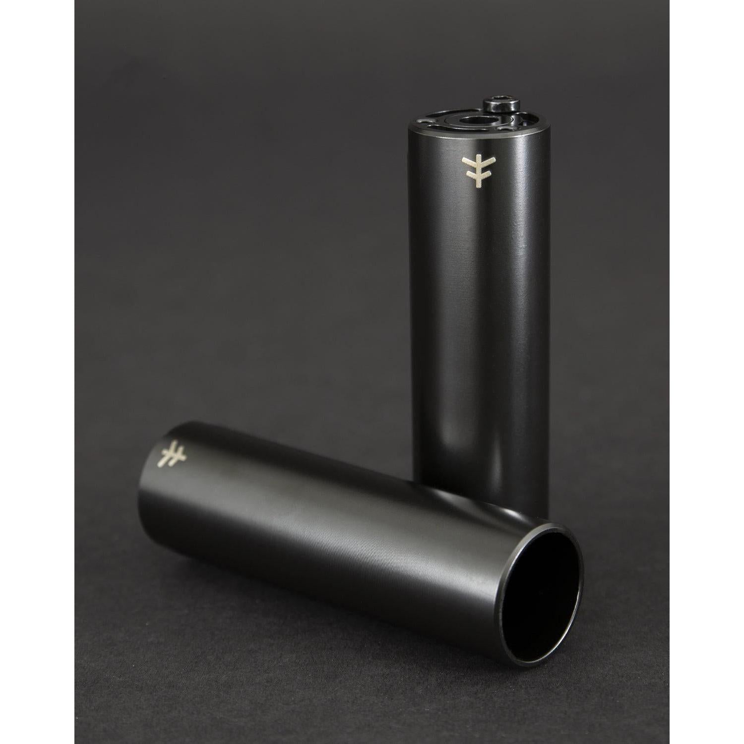 Two Fly Bikes Acero ST Pegs in black rest on a dark surface; one stands upright, showcasing its sleek design reminiscent of CNC machined 4140 Cr-Mo precision, while the other lies down with an air of sophistication.
