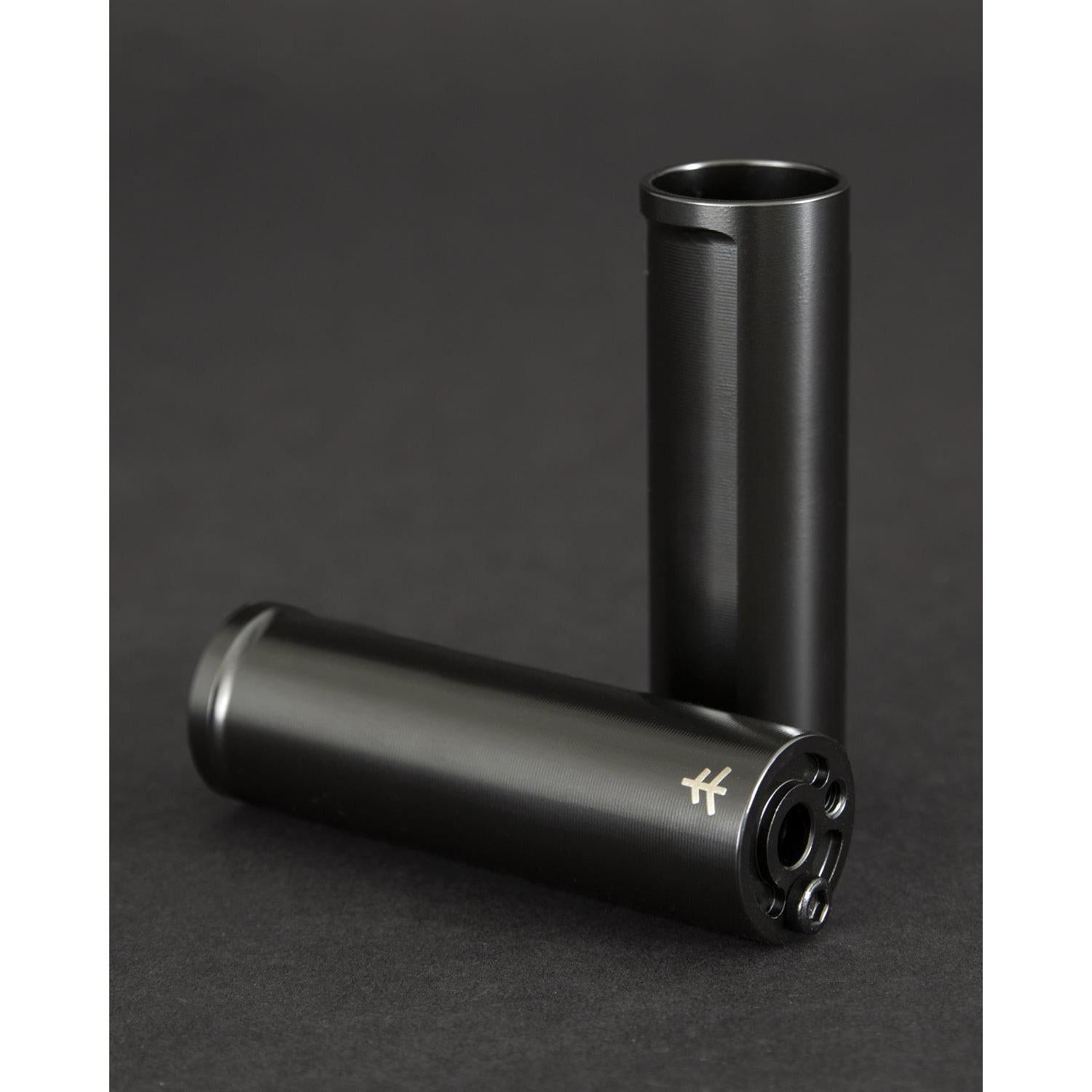 Against a dark backdrop, two cylindrical black metal objects made from CNC Machined 4140 Cr-Mo stand out. One is upright while the other is horizontal, both displaying the Fly Bikes Acero TR Peg logo on one end.
