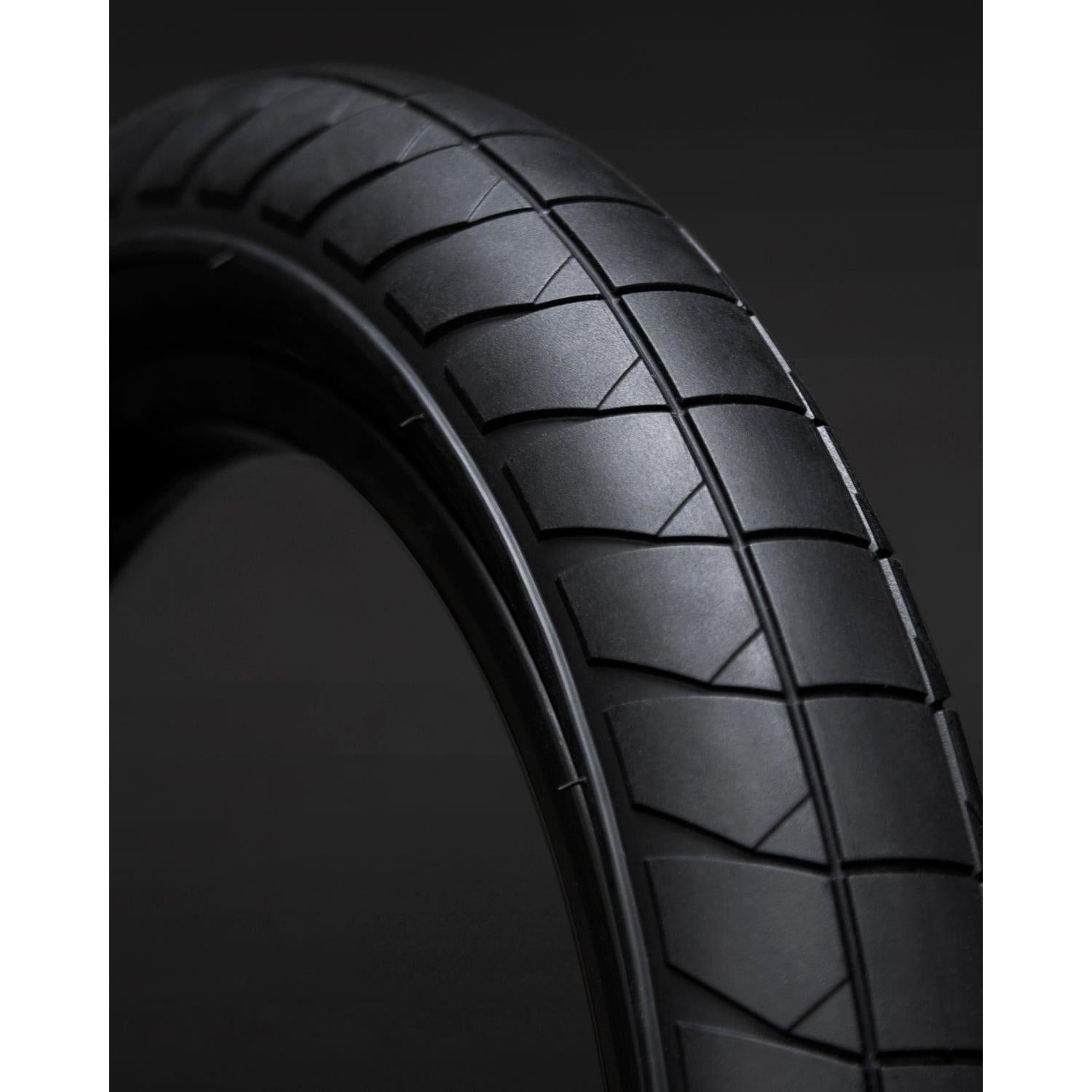 Close-up of the Fly Bikes Fuego Tyre, a black street tire with a geometric tread pattern, showcased on a dark background.