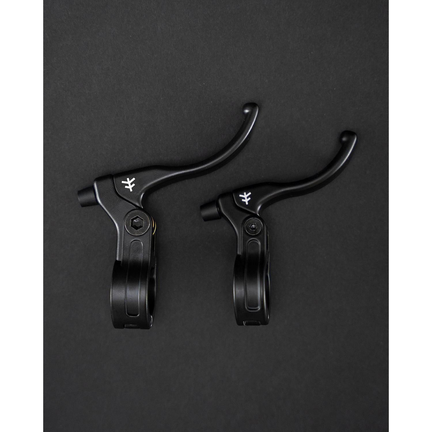On a dark background, two black Fly Bikes Manual Lever Mini BMX brake levers feature a simple white logo and premium CNC brass bushings for smooth operation.