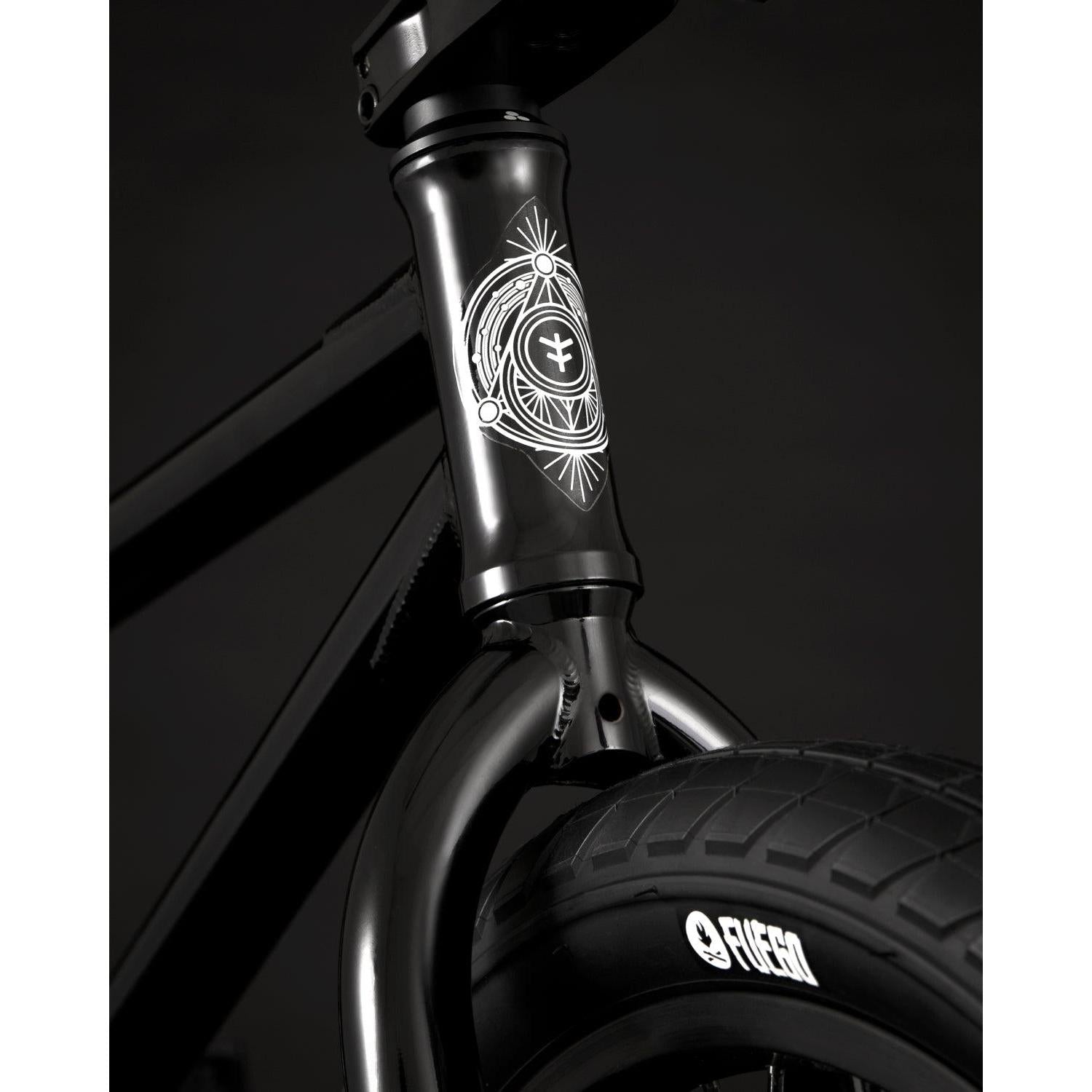 Close-up of the Fly Bikes Proton FC 20 Inch Bike, showcasing its black BMX design with a decorative emblem on the front. The bike features branded "Fiction" tires for added style. Its CrMo frame enhances durability, and the sealed hubs guarantee smooth rides against the dark backdrop.