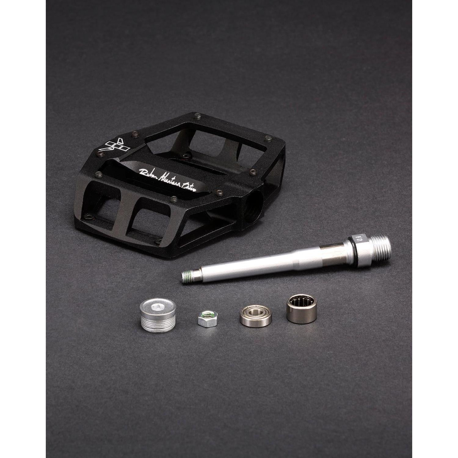 The black Fly Bikes Ruben Aluminum pedal is disassembled, revealing its components: an aluminum pedal body, CNC Chromoly axle, precision bearings, and hardware set against a dark background.