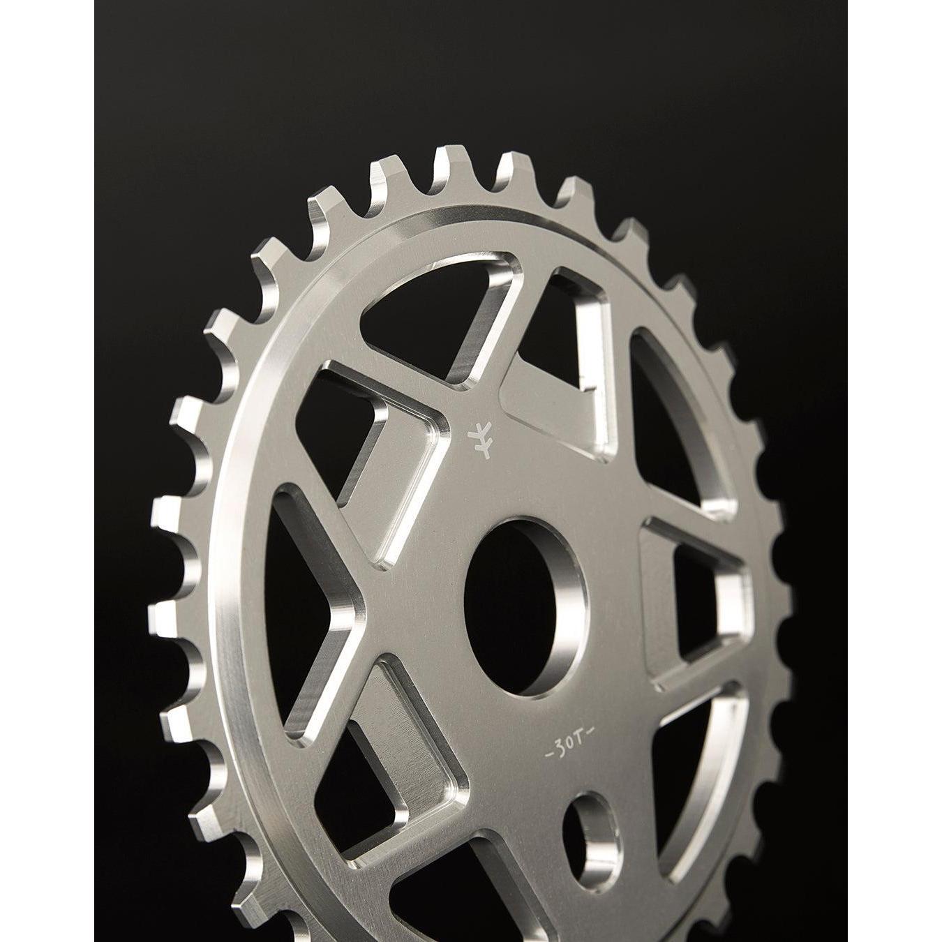 A close-up of the Fly Bikes Tractor TT Sprocket, crafted from durable silver 6061 T-6 Aluminum with multiple teeth and a circular cutout in the center, set against a sleek black background.