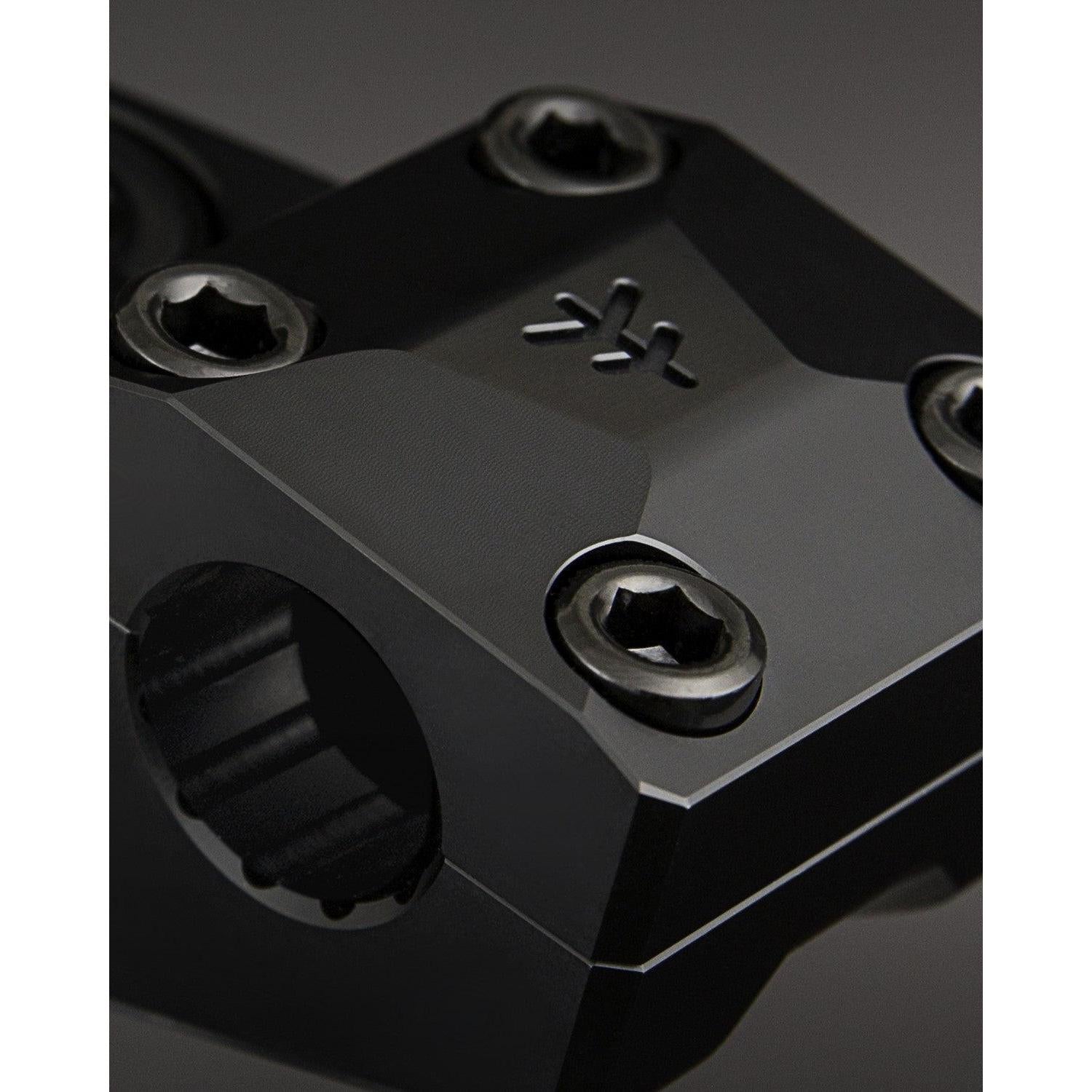 Close-up of a CNC machined black metal component, likely the Fly Bikes Volcano 25 Stem (S), showcasing four screws and a geometric design with engraved initials on top.