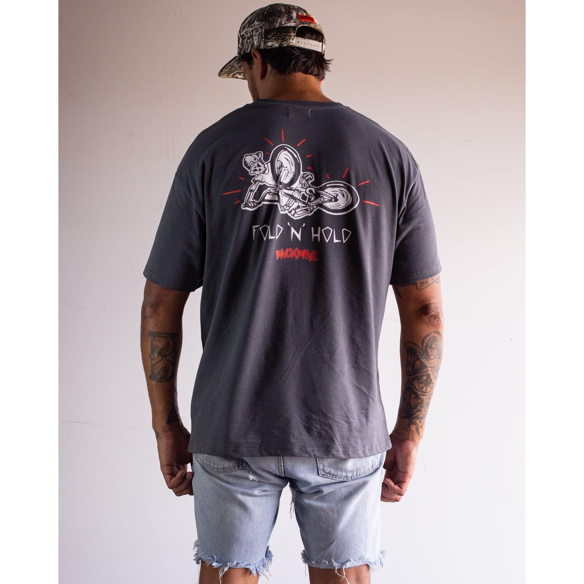 A man in a camo cap, light denim shorts, and a dark Backbone Fold 'N' Hold t-Shirt with a motorcycle graphic stands with his back to the camera.