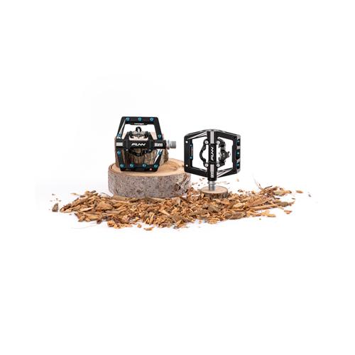 Displayed on a wooden log with wood chips, the Funn Mamba S SPD Pedals in black with blue accents feature a Shimano SPD mechanism, providing precision and reliability ideal for enduro racing.