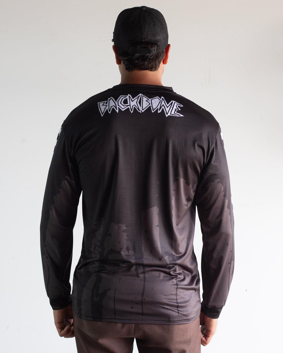 A person faces away, wearing the black Backbone 2026 Gnar Drip Jersey and black cap; bold white "BACKBONE" lettering stands out on the upper back.