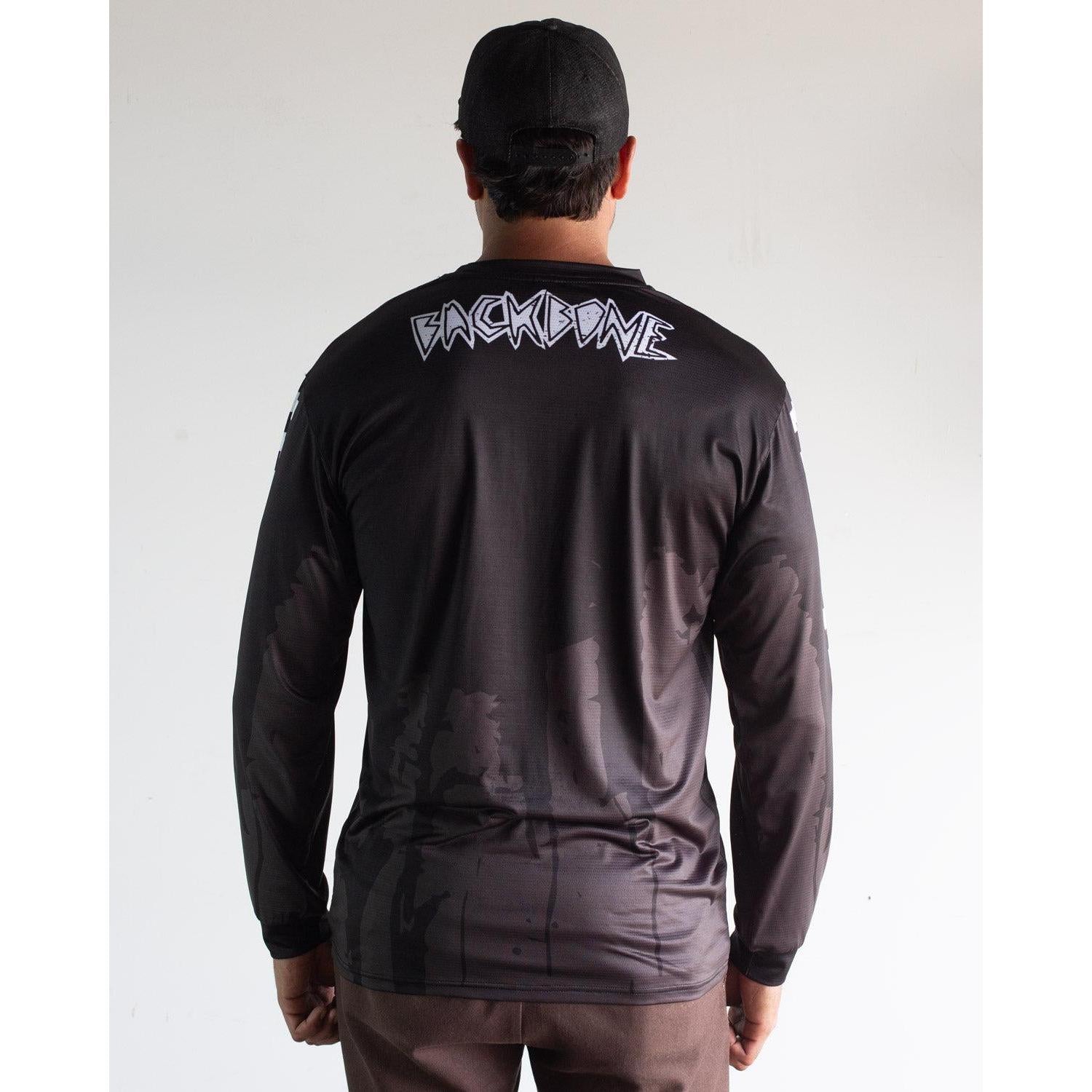 A person faces away, wearing the black Backbone 2026 Gnar Drip Jersey and black cap; bold white "BACKBONE" lettering stands out on the upper back.