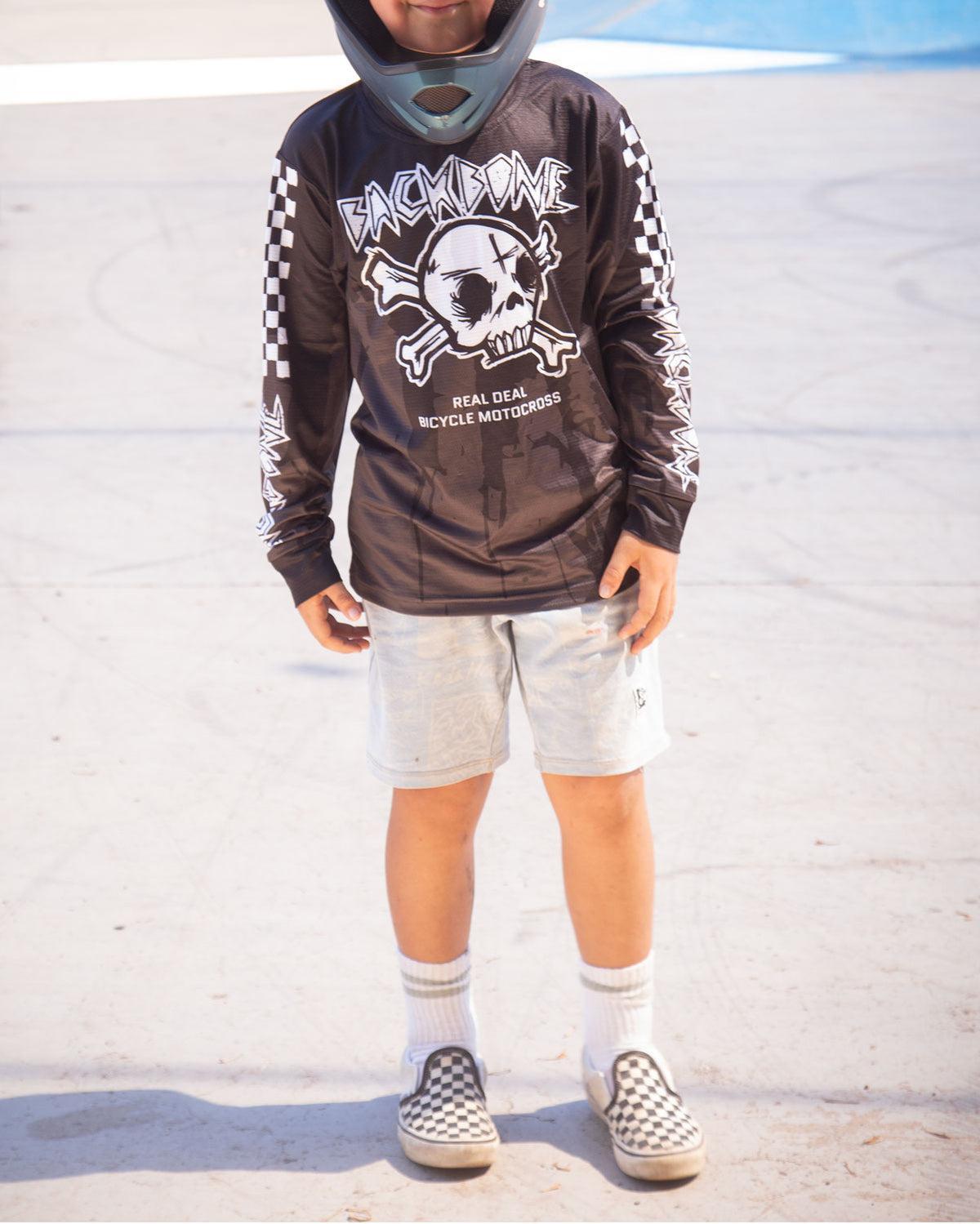 A child in a helmet, Backbone 2026 Gnar Drip Jersey with skull graphic, light shorts, white socks, and checkered slip-on shoes stands on a concrete surface.